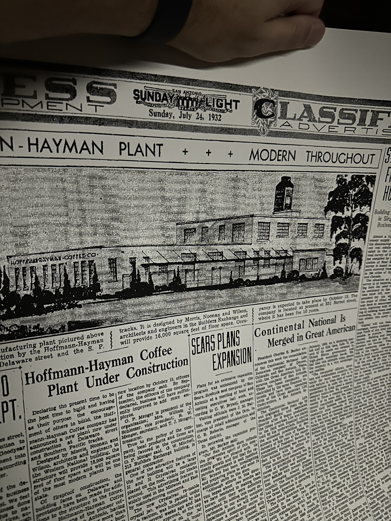 Large-format modern reproduction reprint of a page from the San Antonio Sunday Light of Sunday, 24 July 1932, photographed lying flat on 19 February 2023 — the masthead reading 'SUNDAY LIGHT / Sunday, July 24, 1932', the banner headline 'HOFFMANN-HAYMAN PLANT MODERN THROUGHOUT', a large halftone architectural illustration of the new 1932 factory showing the full-length elevation with its central coffee-silo headhouse tower and row of tall steel-sash industrial windows and 'HOFFMANN-HAYMAN COFFEE CO.' signage across the facade, and the feature article 'Hoffmann-Hayman Coffee Plant Under Construction' by Morris, Noonan and Wilson, 16,000 square feet of fireproof reinforced-concrete construction at Delaware Street and the Southern Pacific tracks, with the 1932 officers listed (G. P. Menger president, Mrs. William J. Schlosser VP, R. W. Menger secretary, T. J. Menger treasurer), and adjacent unrelated articles 'SEARS PLANS EXPANSION' and 'Continental National Is Merged in Great American' on the same page.