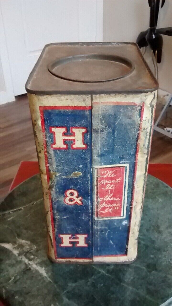 Color photograph documented 2023-05-20 of a heavily worn large square-cross-section bulk-size H and H Blend Coffee tin standing on a green marble side table — viewed at an oblique angle so two faces are visible at once: at left, a narrow side panel carrying a tall vertical 'H & H' monogram in cream-bordered red letters stacked top-to-bottom on a deep blue field; at right, the broader front face showing the early Hoffmann-Hayman slogan 'We roast It / others praise It' in cream lettering inside an ornate red rectangular cartouche; the unpainted flat square top above shows a circular recessed lid well at center with the original press-fit lid missing, the bare metal heavily oxidized.