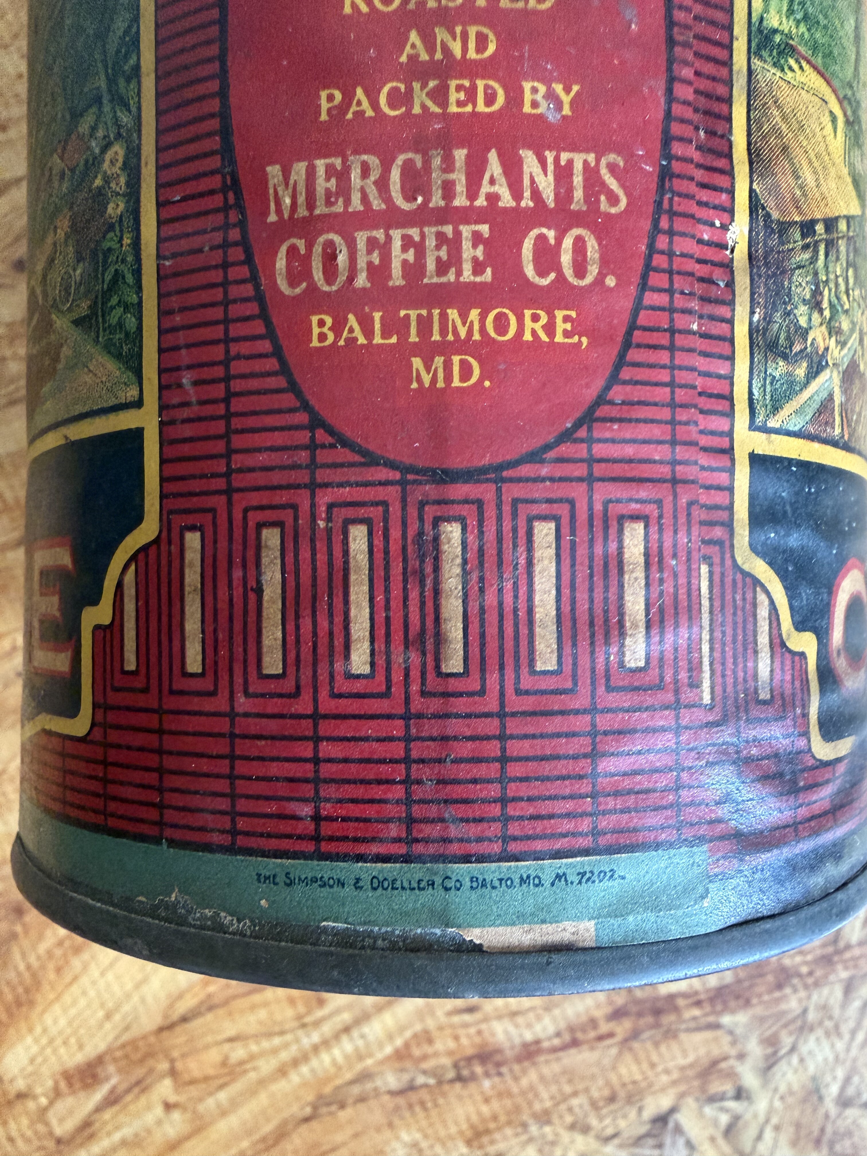 Closeup of the back panel: 'Roasted and Packed By Merchants Coffee Co., Baltimore, MD.' inside an ornate gold-edged oval medallion floating on a red field with vertical-stripe radiator scoring, with the maker's mark 'THE SIMPSON & DOELLER CO BALTO MD M 7202' running along the bottom seam in fine gold capitals