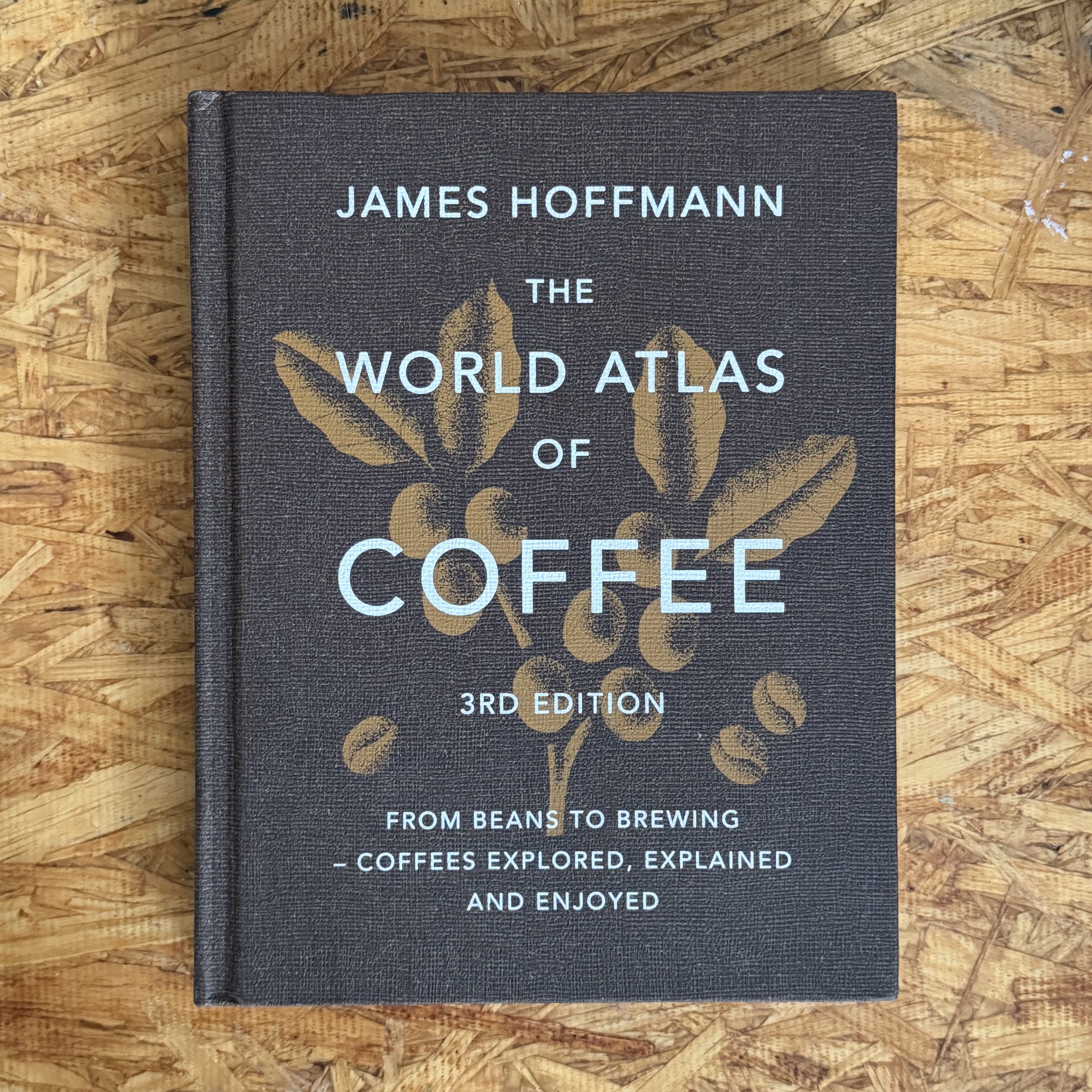 The World Atlas of Coffee 3rd Edition by James Hoffmann — dark charcoal hardcover with gold foil coffee plant and bean illustration, photographed 2026-06-06