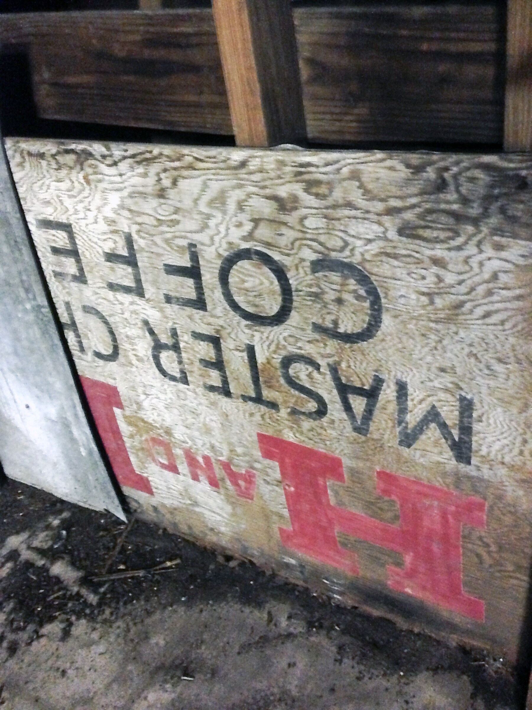 Printed Master Chef plywood still nailed into an interior wall of the east-wing shed, upside down — black COFFEE and MASTER CHEF lines and red H AND H-style lettering inverted beneath wooden beams