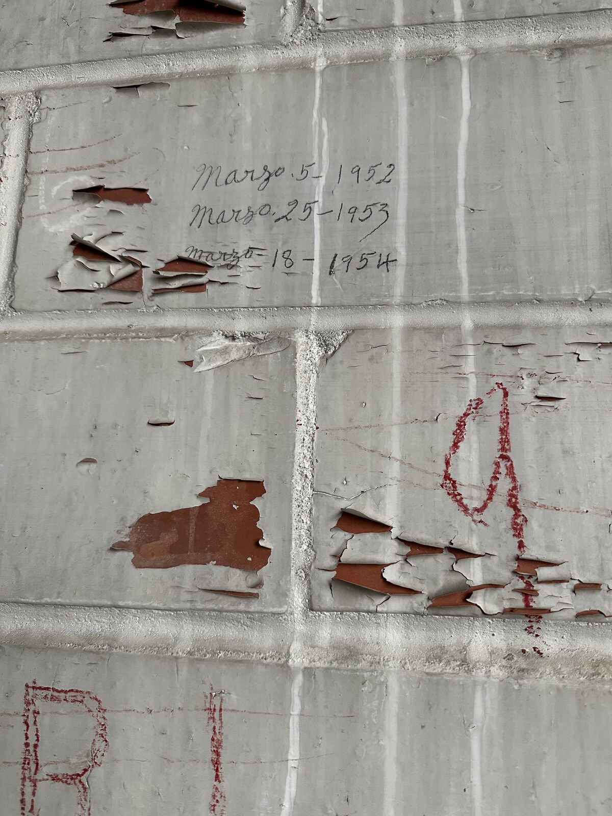 Close-up of the east interior wall of the second-floor expansion room above the office at the Hoffmann-Hayman factory on 1 February 2023 — scored white-painted plaster imitating brick coursing, the paint delaminating to expose warm red substrate underneath, three cursive graphite pencil inscriptions centered in the upper field reading "Marzo 5 - 1952", "Marzo 25 - 1953", and "Marzo 18 - 1954" in the same hand, with red crayon markings and partial red letterforms in the lower half.