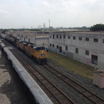 **8 May 2014** (**Apple iPhone 5**) — elevated view from the Hoffmann-Hayman **Delaware Street** context: **Union Pacific** road power (**8809** visible) with **tank cars** on the **industrial lead** beside long **concrete** warehouse façades; flat overcast light, gravel and weeds at grade.