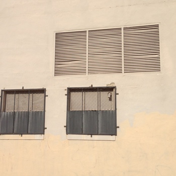 **9 June 2014** (**Apple iPhone 5**) — **south- or side-elevation** detail at Hoffmann-Hayman: wide **horizontal louver**, two **small square** windows with **barred** upper lights and ribbed metal lower closures; stucco **patching** in warm tan on the lower wall, hard sun from camera-right.
