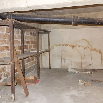 **5 October 2014** (**Sony DSC-RX100**) — low **basement or service** corner at Hoffmann-Hayman: rough **concrete** floor and slab ceiling, **light brick** on one return, **water-streaked** plaster on the back wall, weathered **wood shelving** with a **cardboard** sheet beneath, and a heavy **black cast-iron** building drain or waste line with **PIPE CO** legend on the barrel, elbowing down toward the floor — **same-date** companion to other **vault-zone** infrastructure frames in this gallery.