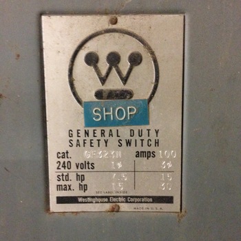 Sheet-metal enclosure on the **first floor** of the Hoffmann-Hayman factory near the older **main-line breaker panels** (the same electrical run documented on the first-floor **[Trumbull Electric breaker-panel circuit directory](/assets/images/gallery/breaker-panel-labels.jpg)**) — angled close-up of the manufacturer nameplate bolted to a grey painted steel knockout box: the circular **Westinghouse** 'W' logo at top (with a small blue rectangular sticker reading **SHOP** in white capitals affixed across the logo), embossed lines reading **GENERAL DUTY SAFETY SWITCH**, stamped catalog **cat. GF323N**, stamped **amps 100**, printed **volts 240**, a molded horsepower rating table (**std.** and **max.** horsepower for single- and three-phase service), tiny line reading **SEE LABEL INSIDE**, and a bottom black banner with white lettering **Westinghouse Electric Corporation** and **MADE IN U. S. A.** at the lower right — surface rust ringing the mounting screws on the brushed-grey housing; photographed **1 December 2014** (iPhone 5)