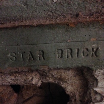 Low-light close-up on the **interior north wall** of the **first floor** at the Hoffmann-Hayman factory — a single masonry stretcher bearing the raised embossed legend **STAR BRICK** in serif capitals (the manufacturer's name-mark block), olive-grey porous clay with a faint horizontal frog line bisecting the lettering, seated in granular buff-grey mortar joints; the wythe sits below a coarse dark relieving course at the top edge of frame and drops into badly spalled and missing mortar toward the bottom, revealing rough subsurface masonry and cavities in deep shadow — readable **maker lettering** tying the site's summary language about **star-stamped San Antonio street bricks** to **in situ masonry** inside the factory's north **first-floor** interior (**interior north wall** per site documentation — **this gallery** places the frame immediately **after** the **[north-end freight elevator look-up](/assets/images/gallery/factory-downstairs-elevator-shaft.jpg)** listing); photographed **4 December 2014** (**iPhone 5**) with grain characteristic of handheld low-light capture