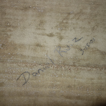 Close-up of the same board-formed reinforced-concrete surface in the vault-adjacent space at the Hoffmann-Hayman factory, photographed 5 October 2014 (Sony DSC-RX100) — seconds after the WATCH YOUR HEAD frame — showing horizontal formwork grain, light tan-grey paste with fine pitting and white speckle, and a handwritten inscription in dark graphite or ink reading the name **Daniel Ruiz** on a diagonal above a short date line **3-15-01** (most naturally read as 15 March 2001 in U.S. month-day-year order), a worker or visitor mark on the 1930s cast concrete