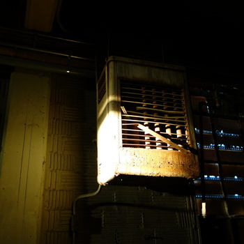 Dim **first-floor**, **north-side** plant-services wall at the Hoffmann-Hayman factory — **gas-fired sectional cabinet heater** (**unit heater**) mounted **high** on vertically coursed **hollow clay tile or structural tile** masonry (regular horizontal recessed rib shadowing typical of glazed structural block), with a plaster-smooth structural **concrete pier** at extreme left intercepting grazing light; the heater's beige/grey **sheet-steel casing** carries a **louver-grille** front (**one bent slat hung down**) over visible **parallel vertical heat-exchanger flues or tubes**, heavy **rust blistering** the lower fascia and sill, service piping or conduit dropping along the masonry below the burner box; darker right field resolves to bolted diamond **steel security grille** across an opening (**daylight-blue** specks visible through gaps); **spatial context**: same **main-line electrical bay** documented on **[Trumbull Electric](/assets/images/gallery/breaker-panel-labels.jpg)** and the **[Westinghouse GF323N safety-switch nameplate](/assets/images/gallery/2014-12-01-factory-first-floor-westinghouse-gf323n-safety-switch-nameplate.jpg)** on the north run — photographed **19 February 2015** (**Sony DSC-RX100**) in high-contrast available light (**strong side falloff**, deep shadow wrapping the masonry field)