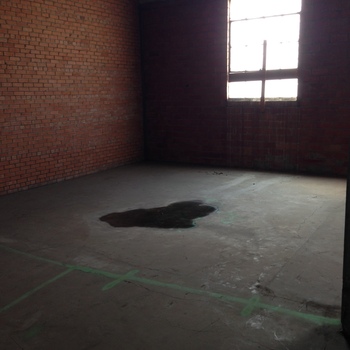 **Second floor**, **northeast wing** (post–1932 **expansion envelope**), Hoffmann-Hayman factory — wide room shot **looking southeast** toward a **tall glazed steel-sash bay** that throws hard **backlit daylight**, leaving **deep shadow** fill on the nearer **warm orange brick wythe** (vertical corner junction at camera-left climbing up to the slab line) while the farther wall washes out silhouette; overhead, **parallel pan-formed reinforced-concrete joist ribs** march across (**corrugated steel-pan bays** readable between them), framing the unfinished **cold shell**; **smooth grey concrete slab** runs underfoot bearing **neon green aerosol/construction-marker** layout arcs and tic marks (**future partition staking**) and a sizable dark **wet pool** pooled mid-slab (**water ingress or condensation**); photographed **13 April 2015** (**iPhone 5**) in the **same upstairs expansion archaeology** corpus as **[Marzo pencil dates](/assets/images/gallery/factory-upstairs-east-wall-marzo-dates.jpg)** **on the expansion east wall**, the **[junction of joist ceiling and original 1932 exterior face](/assets/images/gallery/factory-upstairs-ceiling-febrero-date.jpg)**, **[hole through masonry](/assets/images/gallery/factory-upstairs-expansion-hole-through-original-exterior.jpg)**; related **[Delaware-Street factory profile](/{{ site.baseurl }}/sunday-light-july-1932-factory-article-reprint/)** (**1932** *Sunday Light*, same footprint)