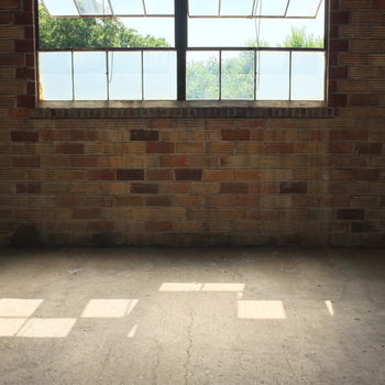 **1 August 2015** (**Fujifilm FinePix X100**) — empty bay: **variegated brick** wall centered on a **twelve-pane steel sash** with **awning** top row open; hard **sun rectangles** on **concrete slab** — quiet interior daylight study at Hoffmann-Hayman.