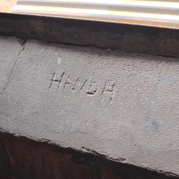 **1 August 2015** (**Fujifilm FinePix X100**) — **hand-scratched H AND H** in a **grey concrete sill** or cap above **dark brick** at Hoffmann-Hayman — informal **company initials** in the architectural fabric.