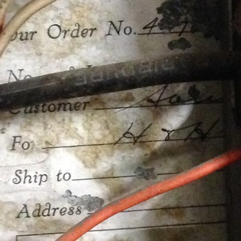 **First floor** electrical **switchgear / breaker-bay run** — extreme close-up of a **yellowed**, **water-and-rust stained** paper slip (likely a shipping or reorder tab) taped or tucked **inside a power-panel enclosure**, photographed **25 November 2015** (**iPhone 5**) — printed form rules read **'Your Order No.'** with handwritten **'44.'**, **'Customer'** with inked cursive **partially obscured** behind a thick **horizontal black insulated service conductor**, **'For'** with clear inked **‘H & H’** tying the paperwork to Hoffmann‑Hayman, blank **Ship to** / **Address** lines beneath; peeling paper and granular grey residue occupy the margins; foreground **conductors** (heavy **black**, bright **orange** thermoplastic, finer **cream** wire crossing upper-left) run across shallow depth consistent with cabling inside cabinet — **companion paperwork** rooted in the same **plant main-line bay** summarized on the **[Trumbull Electric inside-of-door circuit directory](/assets/images/gallery/breaker-panel-labels.jpg)** listing roaster/cooler/mill circuits