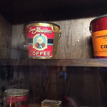 Sam Houston Coffee pail on display at the Witte Museum in San Antonio, photographed through the exhibit glass on 2018-03-24 — red paper label with cream 'Sam Houston' script, an oval portrait of Sam Houston, and 'COFFEE' in block letters on a galvanized pail with wire bail handle; a Longhorn Cafe Coffee tin (San Antonio) sits alongside on the same shelf, with a Red Swan Coffee tin and other small coffee tins on the shelf below