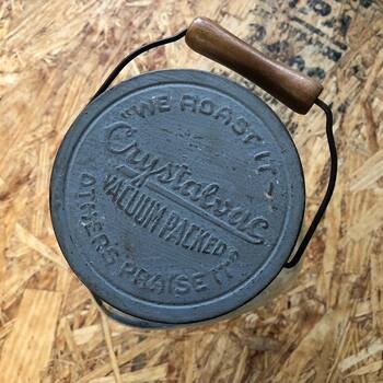 Top-down view of the metal lid: "WE ROAST IT / Crystalvac VACUUM PACKED / OTHERS PRAISE IT" with wire bail and wooden handle