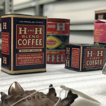 Color photograph **15 October 2019** in the **Witte Museum** collections / study space, San Antonio — white protective foam with a rectangular **H and H Blend Coffee** one-pound **Medium Ground** tin (navy, cream, and red, Roasted and Packed by **Hoffmann-Hayman Coffee Co., San Antonio, Texas**), a smaller **Light Housekeepers Size** **half-pound** H and H Blend tin of the same design family, a large **Family Size** cylindrical H and H tin in red and blue with yellow sunburst visible behind, and at the right edge a **Texas Girl** coffee tin; in the lower-left foreground, the **bottom** of a dark **cast leaf-form ashtray** issued as a **coupon premium** (customers mailed in **H and H** coupons to earn it), showing embossed **'EAT'** / **'SMOKE'** / **'DRINK'** / **'H & H'** / **'COFFEE'** — the same object as the dedicated **KS 193** frame in the 2019 visit post, here with a **white accession string tag**; industrial metal shelving in soft focus behind; **not in the project’s museum collection**