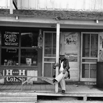 Black-and-white documentary photograph, in the manner of Farm Security Administration / Office of War Information work of the early 1940s, of a rural South Texas Tejano general-store porch — an older mustachioed gentleman in a pale fedora, dark jacket, white shirt, and light trousers sits leaning against a wooden porch post with his left leg crossed over his right; the storefront behind him is densely papered with early-1940s commercial signage including, from left to right, porcelain-enamel 'GARRETT'S Richer and Milder SNUFF' and 'HONEST Mild and Mellow SNUFF' signs and two tall RC Cola bottle-shaped advertising cutouts mounted on porch posts, a hand-lettered Spanish-language 'ESPAUDA Health Club / Conserve su...' window sign, window-display items including cardboard cutouts and a 'LOOK 25¢' magazine, through the door-glass 'UNION LEADER' and 'UNION STANDARD' tobacco posters with Chesterfield and Bugler tobacco ads and a 'RIPPLE' sign, an 'Ask for the best / TINSLEY'S / NATURAL LEAF' tobacco sign on the low porch railing, and — mounted squarely on the porch railing facing the street — a large dark-ground porcelain-enamel **'H AND H COFFEE'** sign whose big white serif 'H AND H' type matches the other H and H Coffee porcelain signs in the project; at the right edge a gravity-feed visible gasoline pump with a small framed photograph affixed to its tank stands in front of the rear fender and running board of a late-1930s automobile, and at the left edge a Spanish-language movie poster reading 'ALAMO / SABADO - DOMINGO / MUJER MEXICANA / ELVA SALCEDO / J.J. MARTINEZ CASADO / MARGARITA CORTES' — the 1942 Miguel Contreras Torres film, dating this photograph to 1942 or shortly after