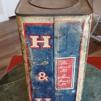 Color photograph documented 2023-05-20 of a heavily worn large square-cross-section bulk-size H and H Blend Coffee tin standing on a green marble side table against a wood-floor and white-door domestic interior — viewed at an oblique three-quarter angle so that two adjacent painted faces are visible at once: at left, a narrow side panel carrying a tall vertical 'H & H' monogram in cream-bordered red letters stacked top-to-bottom on a deep blue field framed by a red double-line border on a cream ground, with the 'H', the ampersand '&', and a second 'H' arranged as three separate red cartouches running down the panel; at right, the broader front face of the tin showing a deep blue field with an ornate red rectangular cartouche bearing in cream script-and-roman lettering the early Hoffmann-Hayman slogan **'We roast It / others praise It'**; the painted surfaces show extensive scratch wear, paint loss along the corner crease, surface scuffing, and dust; the unpainted top of the tin is a flat square plate with a circular recessed lid well at its center (the original press-fit lid is missing), the bare metal heavily oxidized; the same red-white-and-blue palette and slogan as the Euless, TX three-pound H and H Blend tin in the museum collection, but in a substantially larger bulk format — not in the museum collection