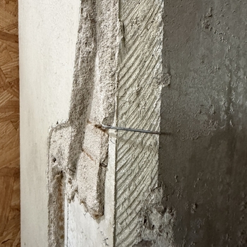 **Hoffmann-Hayman factory**, **first floor**, **bay one** beside the **office** — **column encasement** opened for investigation or repair (per site note; same **first-floor** **column** session as **[companion lath-tear frame](/assets/images/gallery/2026-04-24-factory-first-floor-bay-one-office-column-expanded-metal-lath-protruding.jpg)**) — upright tight photograph of **multi-layer vertical strata** at a **ragged vertical break**, documented **24 April 2026** (**Apple iPhone 17 Pro Max**) — reading left to right: narrow **tan oriented-strand-board (OSB)** edge showing **pressed wood strands**; flat **off-white paint-finish** gypsum or plasterboard skim; jagged **tear** through a **light porous mortar / patch fill** exposing coarse aggregate; adjacent **diagonal-grooved plaster scratch coat** mechanically keyed for bonding; right **massive grey masonry or cast core** with pitted irregular face; **thin steel wall tie** (masonry connector wire) projects horizontally from the textured intermediate zone into the grey mass with a **slightly curved tip** — **contemporary column-wrap forensic** complement to the **January 2025** deliberately opened **[1932 exterior-face cross-section](/assets/images/gallery/factory-upstairs-expansion-hole-through-original-exterior.jpg)** but here foregrounding **modern sheathing and interior finish buildup** over historic mass