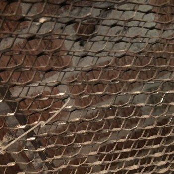 Exposed expanded-diamond metal lath underlying the plaster wall treatment in the first-floor bathroom at the Hoffmann-Hayman factory, immediately adjacent to the vault — with patches of plaster fallen away to reveal the lath and the red brick wall behind it