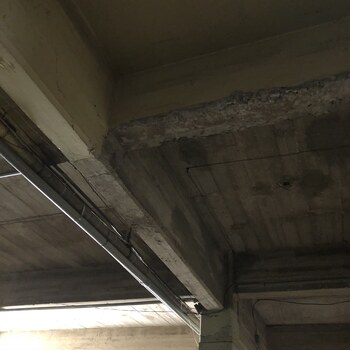 Looking up at a first-floor ceiling corner at the Hoffmann-Hayman factory, where a rough board-formed reinforced-concrete beam meets its column and the slab above — the horizontal grain of the 1930s formwork boards is pressed into the beam's vertical face, a galvanized water line runs along the beam at left, and steel electrical conduit is strung along the beam soffit; exposed aggregate is visible where a shoulder of concrete has spalled away at the beam's top edge