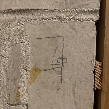 Close-up of an informal pencil sketch drawn in graphite directly on a troweled white-stucco interior wall on the first floor of the Hoffmann-Hayman factory, photographed on 11 March 2022 — at the foot of the stairs up to the second floor, right at the edge of a later-cut secondary doorway into a first-floor bathroom, where the wood jamb and a brass door hinge are visible at the right edge of the frame (with a section of twisted-fibre rope running through the gap at mid-height) and a thickened projecting stucco ledge crosses the top of the frame; the sketch itself is a schematic composition of an irregular pointed shape above a small rectangle with a horizontal line extending right (ending in a short vertical tick) and a vertical line descending below, reading loosely as a rough elevation-style diagram, and a torn-piece-shaped yellow-amber residue of aged masking-tape adhesive is soaked into the plaster at lower-left — a second human trace beside the pencil mark itself