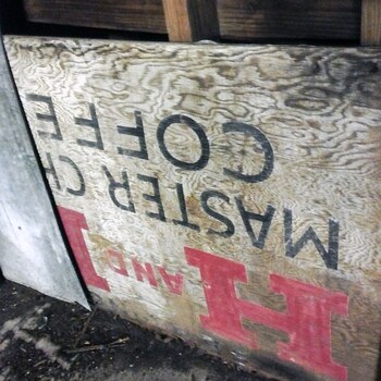 **May 2014** (**iPhone 5**) — **metal-roofed east-wing shed** at Hoffmann-Hayman: **printed Master Chef** plywood used as **interior sheathing**, **upside down** under joists — **COFFEE** / **MASTER CHEF** and **H AND H**-style **red** and **black** art still legible. Documented in [East-wing shed — Master Chef plywood and shipping-crate boards in the walls](/east-wing-shed-salvaged-crate-wood-in-walls/).
