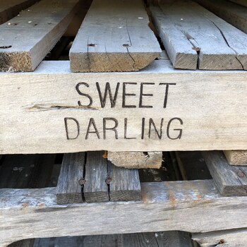 Weathered honey-colored wood crate board stencil-lettered 'SWEET DARLING' in black block caps, resting on a stack of reclaimed pallet and crate wood at the Hoffmann-Hayman factory — Sweet Darling was a mid-twentieth-century produce/fruit shipping brand, and this board is one of the reclaimed pieces kept on the property