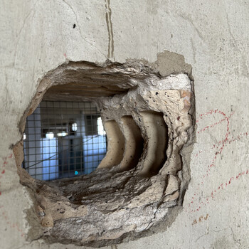 A roughly oval, rounded-irregular **hole punched through the original 1932 exterior face of the plant** as it stands today inside the second-floor expansion room above the office at the Hoffmann-Hayman factory, photographed on 5 January 2025 — the wall in question being the **same exterior masonry face documented in the [adjacent ceiling-junction frame](/assets/images/gallery/factory-upstairs-ceiling-febrero-date.jpg)** with the 'FEBRERO 11 – 53' pencilled joist, now reading as an interior wall because the post-1932 second-floor expansion was built over and beside it; the camera-facing surface is a smooth grey **stucco / cement-render interior finish** with hairline shrinkage cracks tracing through the field and a faint **red crayon / lumber-keel scribble** in the upper right (matching in material and hand the same red crayon marks already documented on the [east wall of this room beside the 1952–54 'Marzo' pencil dates](/assets/images/gallery/factory-upstairs-east-wall-marzo-dates.jpg)); the broken edges of the hole expose the wall in cross-section, revealing what reads as a textbook **early-1930s fire-resistant industrial wall buildup** — a course of warm orange-clay **fired brick veneer** visible at the upper margin of the cut, behind it a stack of three to four characteristic curved/lobed cell profiles consistent with the broken interior webs of **hollow structural clay tile** (terra-cotta hollow tile blocks) used as backup masonry behind the brick veneer, and the smooth grey stucco render skinning the interior face — the kind of brick-veneer-over-hollow-tile composition consistent with the *San Antonio Sunday Light*'s [July 1932 description of the new plant as 'fireproof reinforced-concrete' construction](/sunday-light-july-1932-factory-article-reprint/) (concrete frame plus fireproof masonry infill); through the hole the room beyond is visible — a clear gridded blue-and-silver field reading as one of the building's industrial windows with security mesh / wire-grid in front of it, daylight coming through, the same kind of caged-window run already documented on the [west flank from the rail siding at dusk in February 2019](/assets/images/gallery/factory-south-end-from-tracks-2019.jpg)