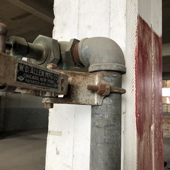 The second of two W. D. Allen Mfg. Co. fire-hose stations documented on the second floor of the Hoffmann-Hayman factory — a cast-iron swing bracket bolted to a white-painted structural column, maker's plate reading 'W. D. ALLEN MFG. CO. — CHICAGO, NEW YORK — PATENTED AUG. 1924', with a threaded elbow feeding a galvanized standpipe running down beside the column and a red painted band on the adjacent column marking the fire-equipment station