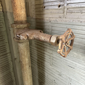 Cast-iron handwheel gas-line shutoff valve in the northwest corner of the second floor of the Hoffmann-Hayman factory, photographed on 5 January 2025 — a heavily oxidized vertical iron riser pipe runs floor-to-ceiling against the wall corner, and a horizontal valve body is clamped to the riser by a bolted bracket (two visible through-bolts gripping the riser) projecting a short horizontal stub out into the room and terminating in a polished brass/bronze hex union nut and a four-spoke cast-iron round handwheel painted rust-red, the union nut sitting between the valve body and the handwheel-side connection so the valve can be unscrewed for service; the assembly is a typical period plant-services branch shutoff sitting in the corner where two whitewashed plaster-over-masonry walls meet, both wall faces carrying a fine, regular horizontal ribbing/banding texture (consistent with either scored scratch-coat plaster or impressions of underlying lath showing through thin whitewash) with a patch of bare red-brick masonry exposed in the upper right corner where the whitewash has worn or fallen away — same kind of whitewashed-brick second-floor interior surface that surrounds the building's other upstairs plant-services hardware