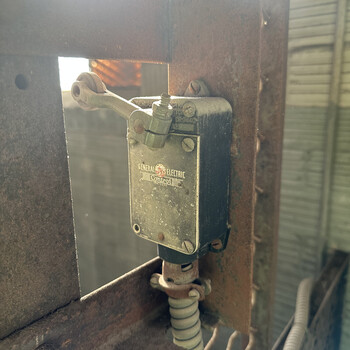 Heavily oxidized **General Electric 'Control'-branded roller-lever travel limit switch** mounted on the riveted steel structural frame of the same smaller second lift at the Hoffmann-Hayman factory — the smaller mechanism (distinct from the main interior freight elevator) fitted with the **exterior access door** opening to the **track side** of the building onto the Southern Pacific siding — photographed on 5 January 2025; a small black-painted cast-metal enclosure carries a brass cover plate embossed in raised lettering with 'GENERAL ELECTRIC' across the top in a banner, a circular red-and-white **GE monogram badge** at center, and 'Control' in flowing italic script below the monogram (the standard pre-1960s GE 'Control' nameplate styling); the cover is held on by four slotted machine screws (one at each corner) with two further visible screws on the body, and out of the top of the case projects a steel **roller-lever actuator arm** pivoting on a hex-cap screw with a brass actuating roller at its outer end (positioned to be physically tripped by an end-of-travel cam or lug on the moving lift platform when the lift reaches the top or bottom of its travel, breaking the contacts inside the case and cutting power to the lift motor — a standard travel/end-of-travel limit switch); the bottom of the case is fitted with a threaded conduit hub feeding a length of **white/grey-jacketed flexible BX-style armored power cable** that loops down past additional wire mesh visible at lower-right, and the whole assembly is bolted to a heavy structural steel angle/channel post that's part of the lift's frame, with weathered tongue-and-groove vertical wood siding visible through the gap on the right of the frame and bright sunlight breaking through behind — most likely from the **trackside exterior access door** itself standing open at the moment of the photograph