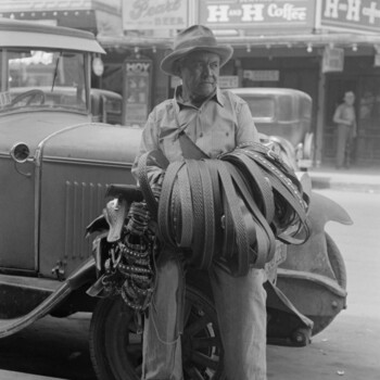 Black-and-white street photograph of a belt peddler in a felt hat leaning against a late-1920s Ford sedan on a San Antonio sidewalk, an armful of tooled leather belts draped over one arm, with storefront signs behind him reading 'Oriental Cafe and Bar — Serving H&H Coffee' and 'H & H Coffee' alongside a Pearl Beer sign